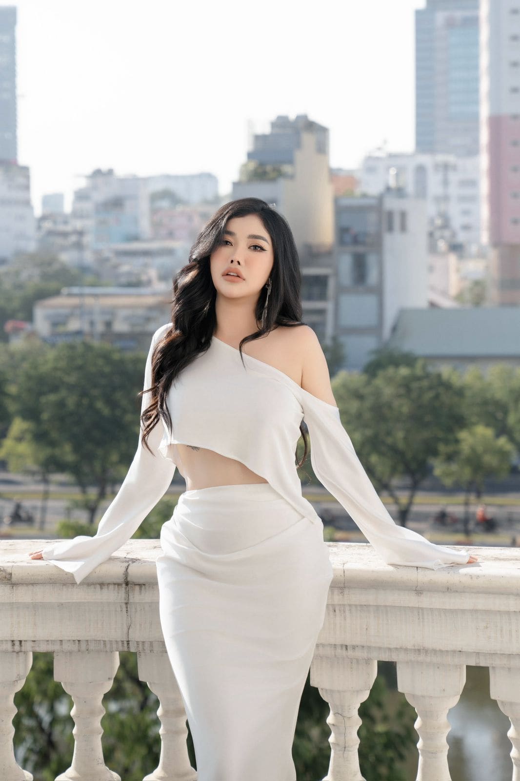A woman in a white one-shoulder cut-out top and long skirt stands by a stone balustrade, framed by a city skyline and trees in the background.