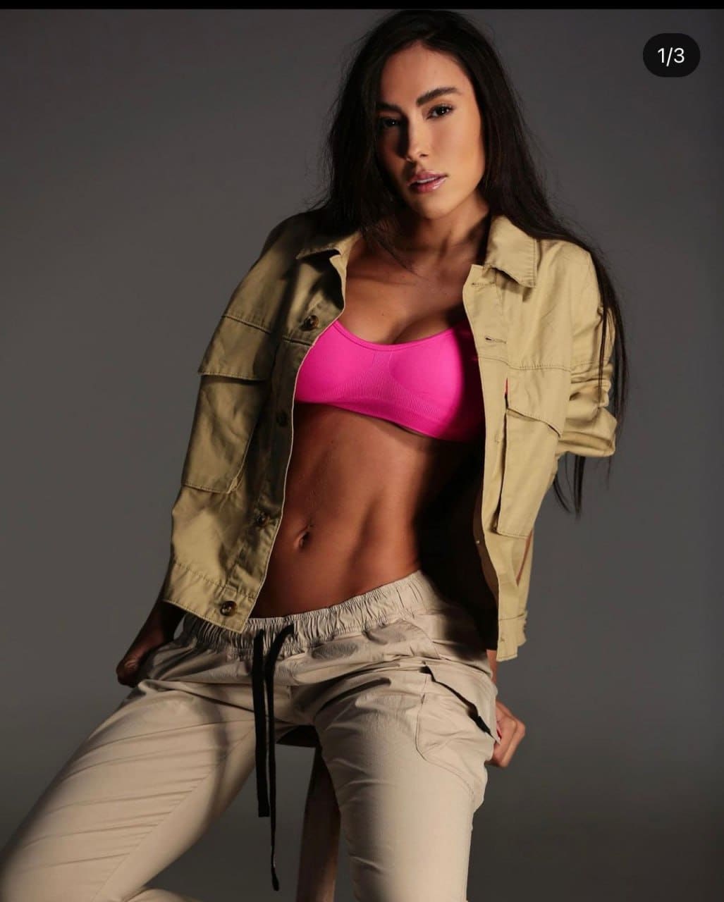 A woman sits on a stool in a studio wearing an open beige jacket over a pink sports bra and light drawstring pants.