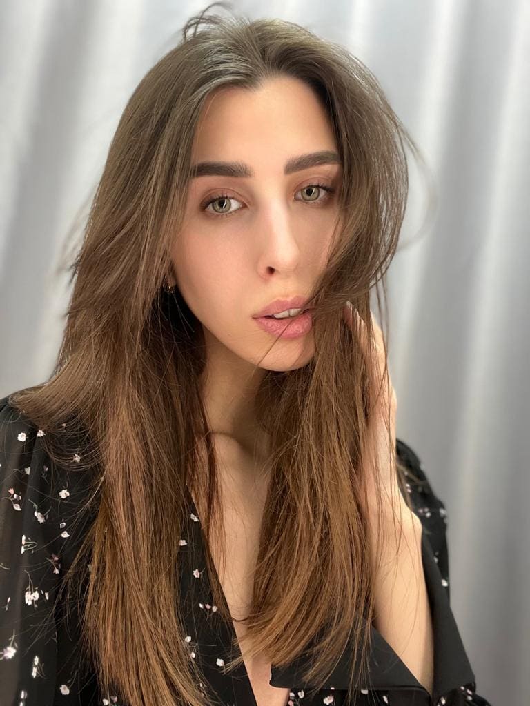 Yana stands before a white curtain, wearing a black floral top with a deep V-neckline and raising one hand near her face.