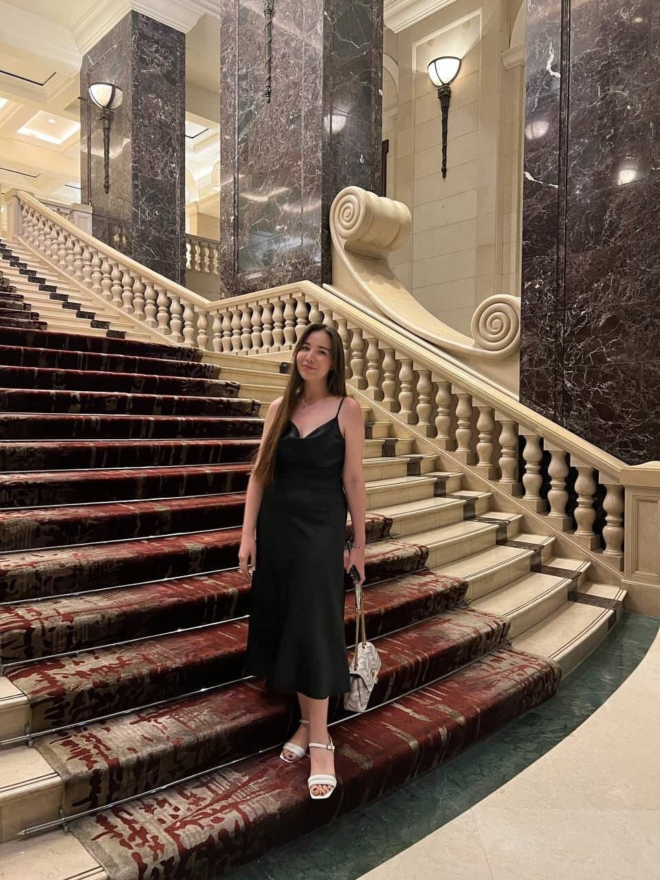 Woman in a black midi dress and white heels stands on a carpeted staircase in a marble hall.