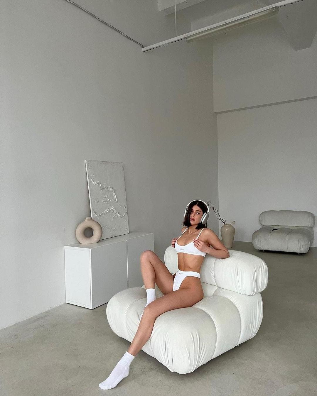 A woman sits on a white sofa in a white lingerie set and socks, wearing headphones and holding the cups of her top.
