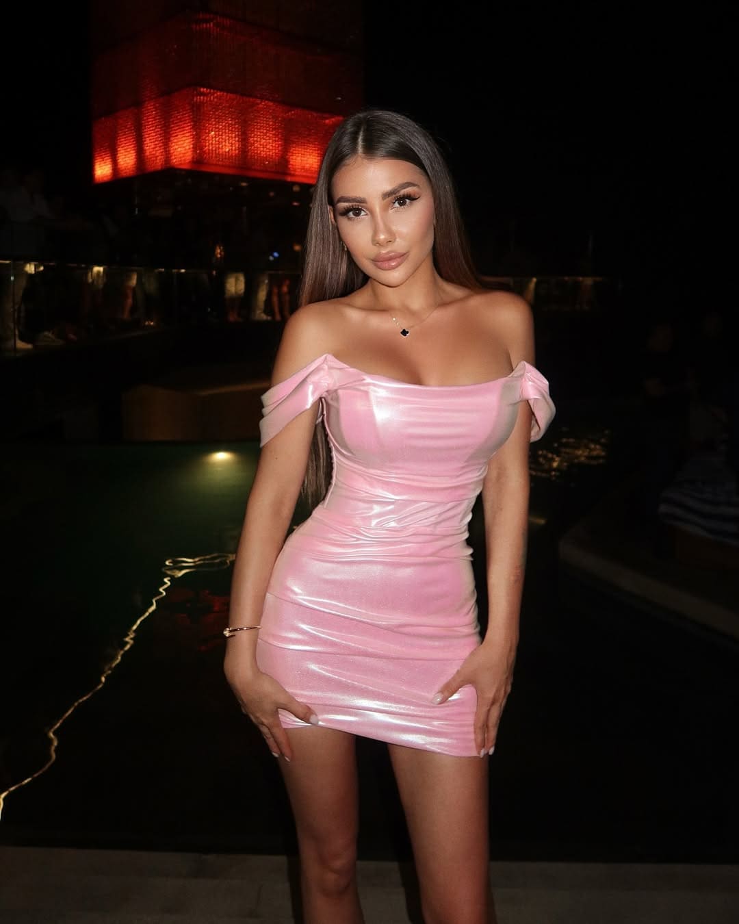 A woman in a pink satin off-the-shoulder mini dress poses with hands on hips against a dark background with red lighting.