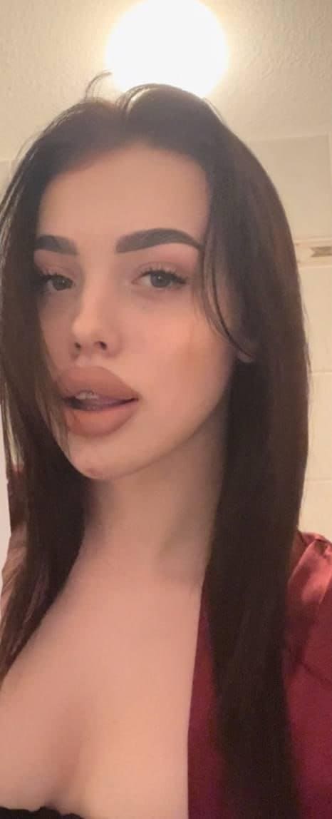 Close-up selfie in a deep-red satin robe over a black strapless top, against a plain wall.