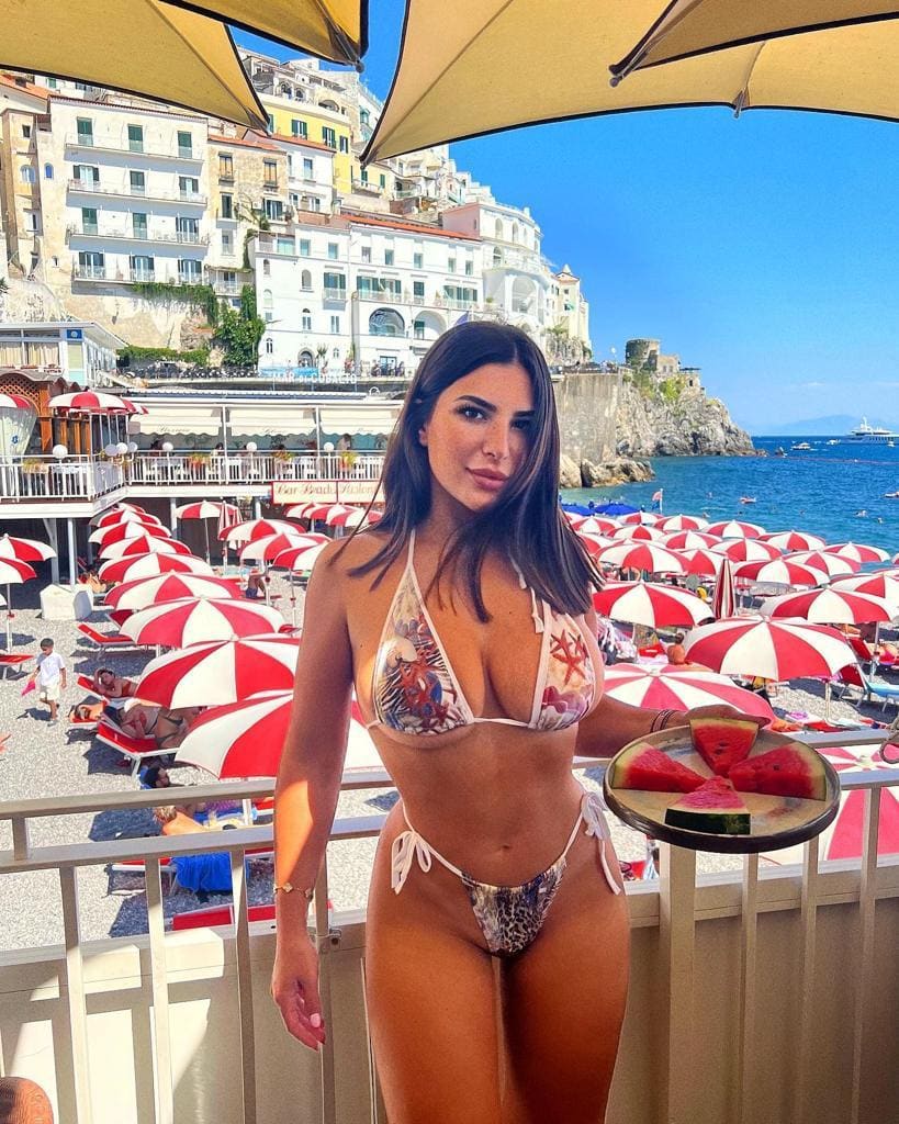 On a balcony, she holds a plate of watermelon in a patterned bikini, overlooking a pebble beach, umbrellas, and a hillside town.