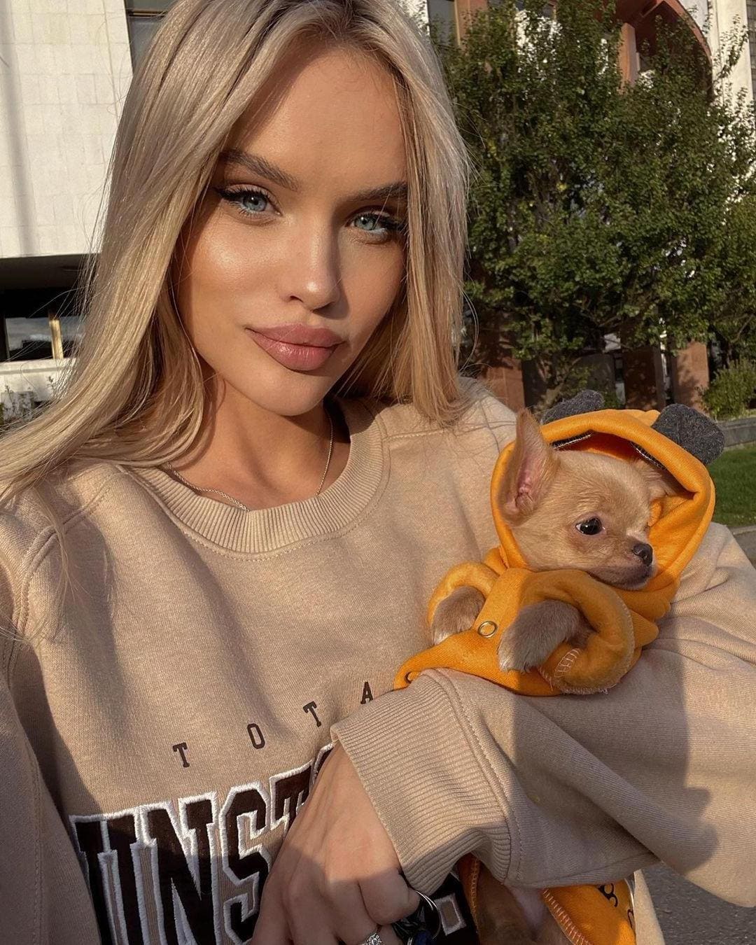 Emma holds a tan puppy in an orange outfit while wearing a beige sweatshirt outdoors near greenery.