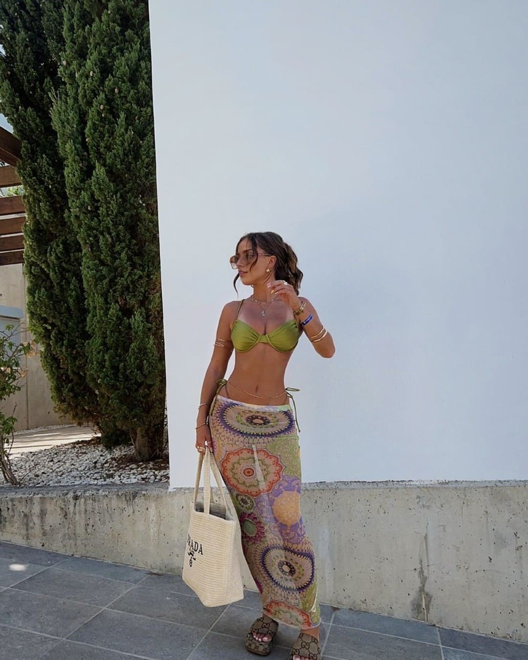 A woman in a green bikini top, patterned skirt, and sunglasses stands in profile against a white wall, holding a woven tote bag.