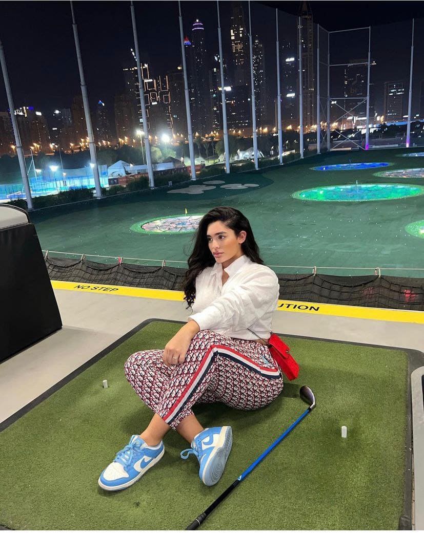 She sits cross-legged on a rooftop driving range at night, wearing a white shirt and patterned pants, with a city skyline behind her.