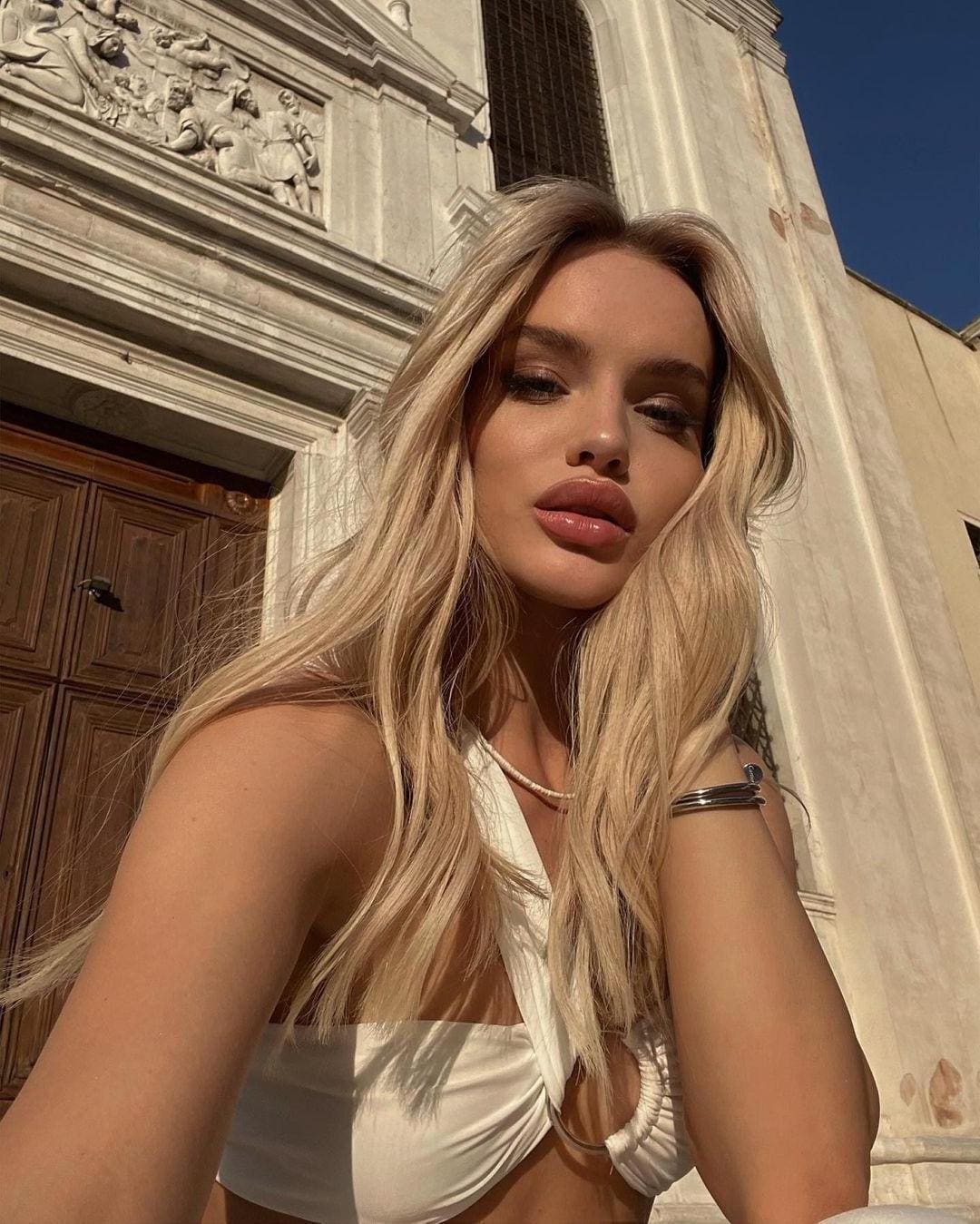 Blonde woman in a white cut-out top poses in front of a carved stone facade with a wooden door.