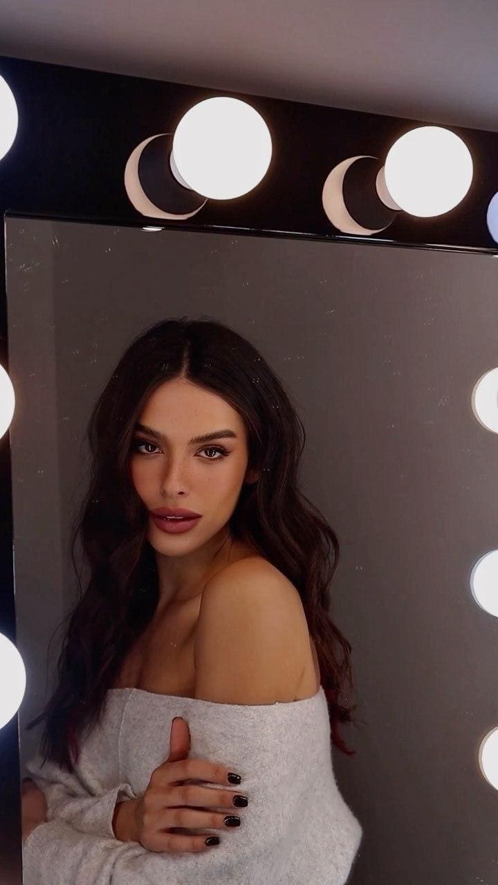 A woman in a grey off-the-shoulder sweater stands before a lighted mirror, her hand resting on her chest with dark nail polish visible.