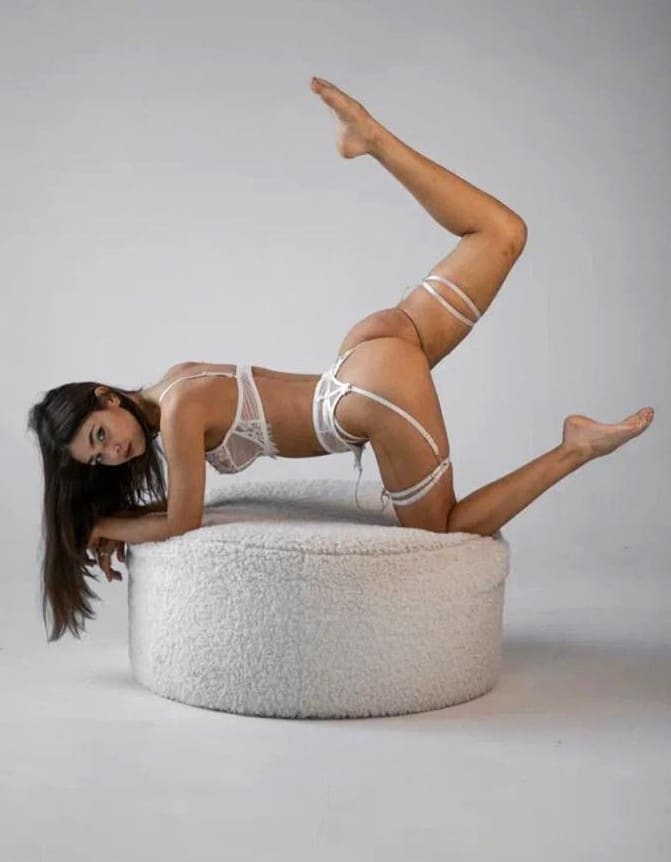 Woman lies on her side on a white fluffy cushion in white lace lingerie, one leg raised, against a light background.