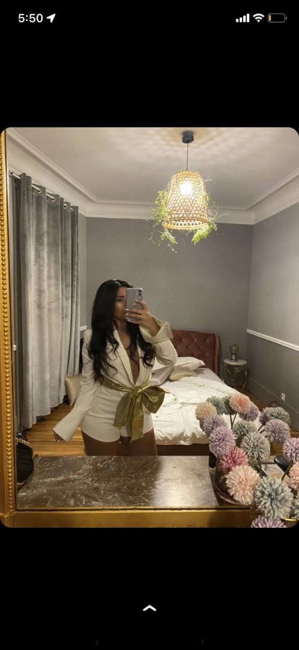 In a bedroom mirror selfie, she wears a white blazer with a gold sash and brown skirt, with a bed and woven lamp visible.
