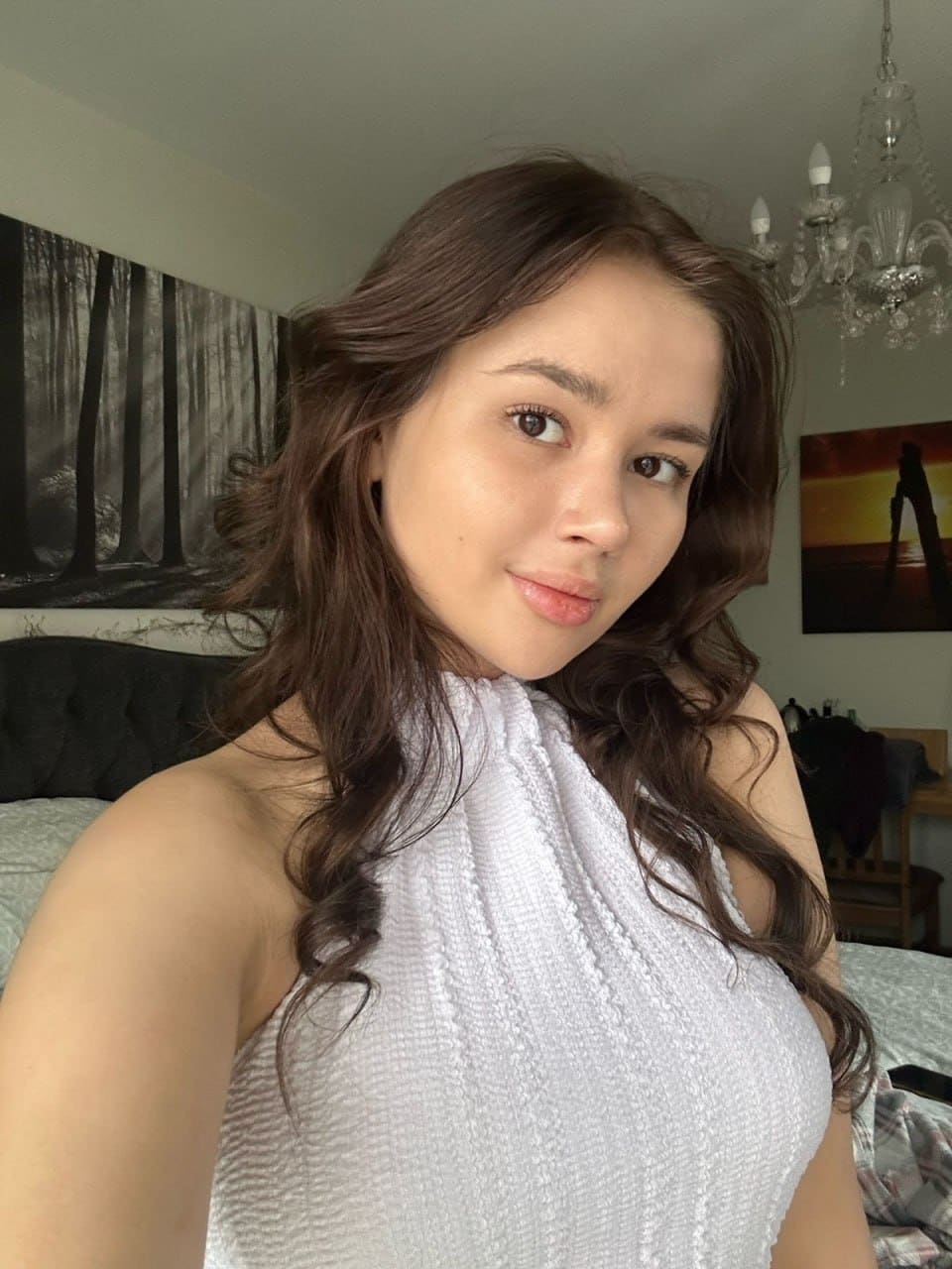 A woman in a white sleeveless halter top stands in a bedroom, with a bed, wall art, and a chandelier visible in the background.