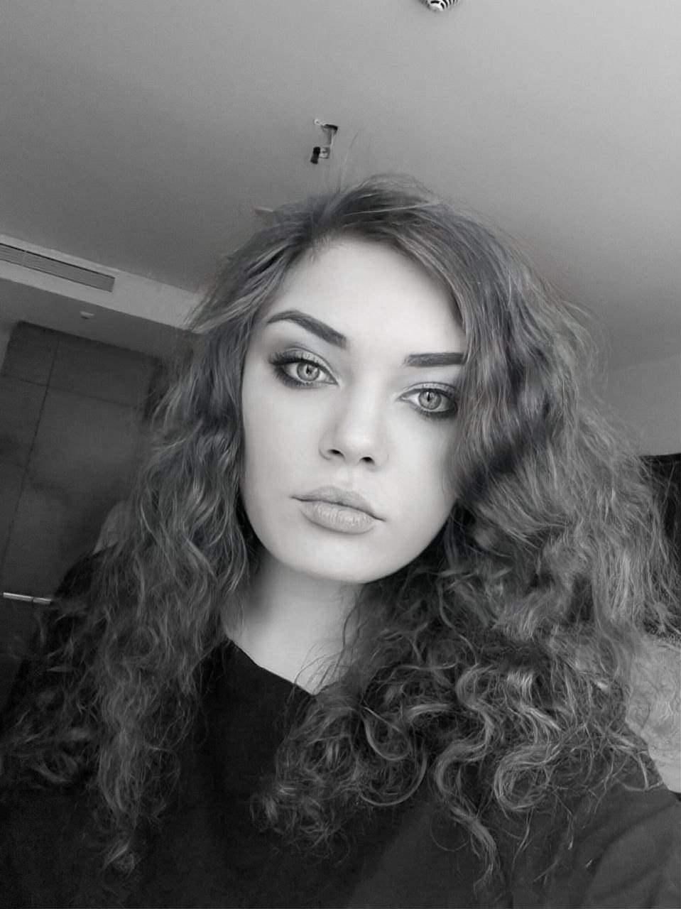 Black-and-white close-up of a woman with curly hair and heavy makeup, low angle, indoors with a ceiling light visible.