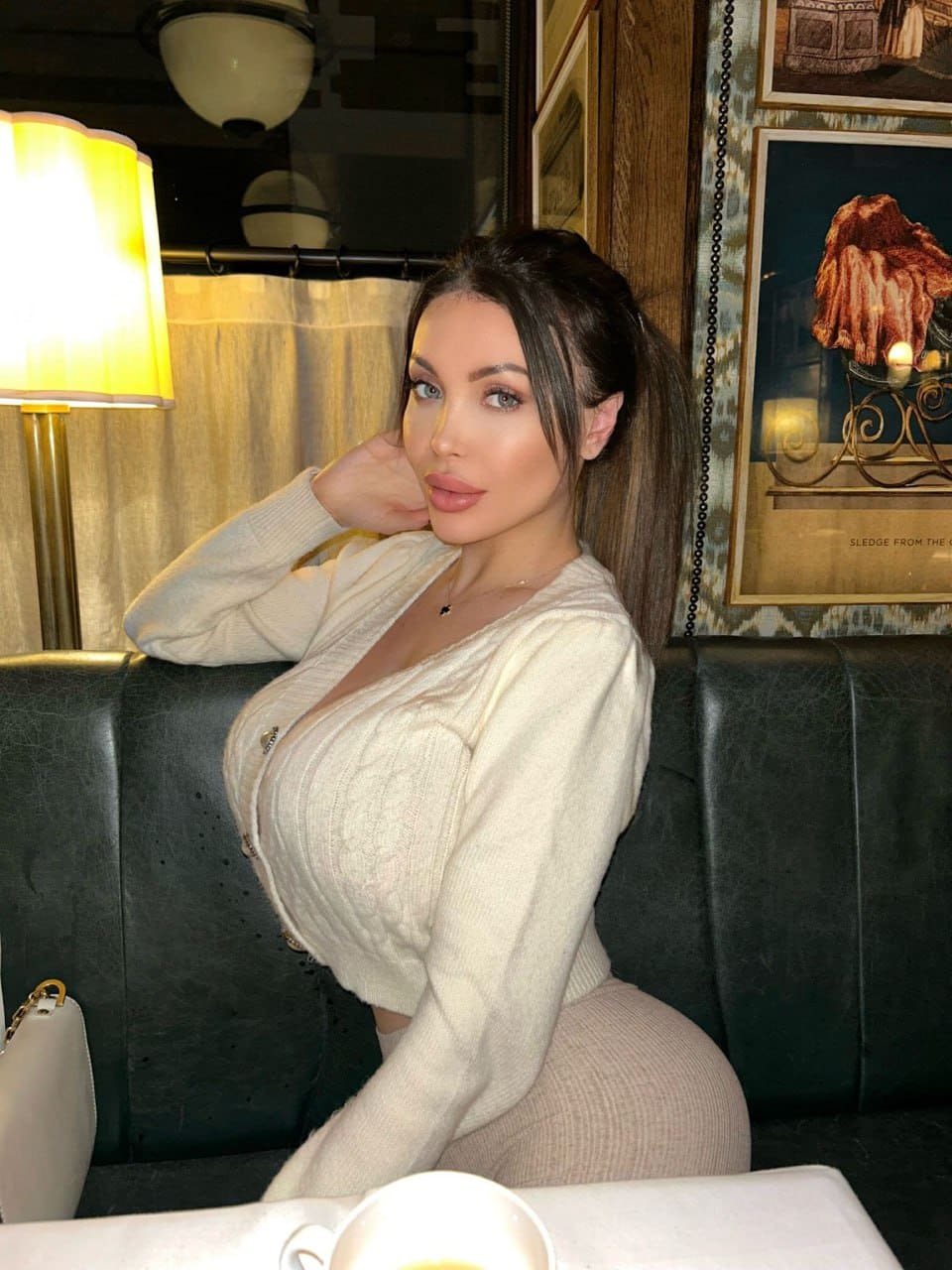 Sitting on a black leather couch, she wears a cream cable-knit cardigan and light pants, resting a hand near her neck in a lamp-lit room.