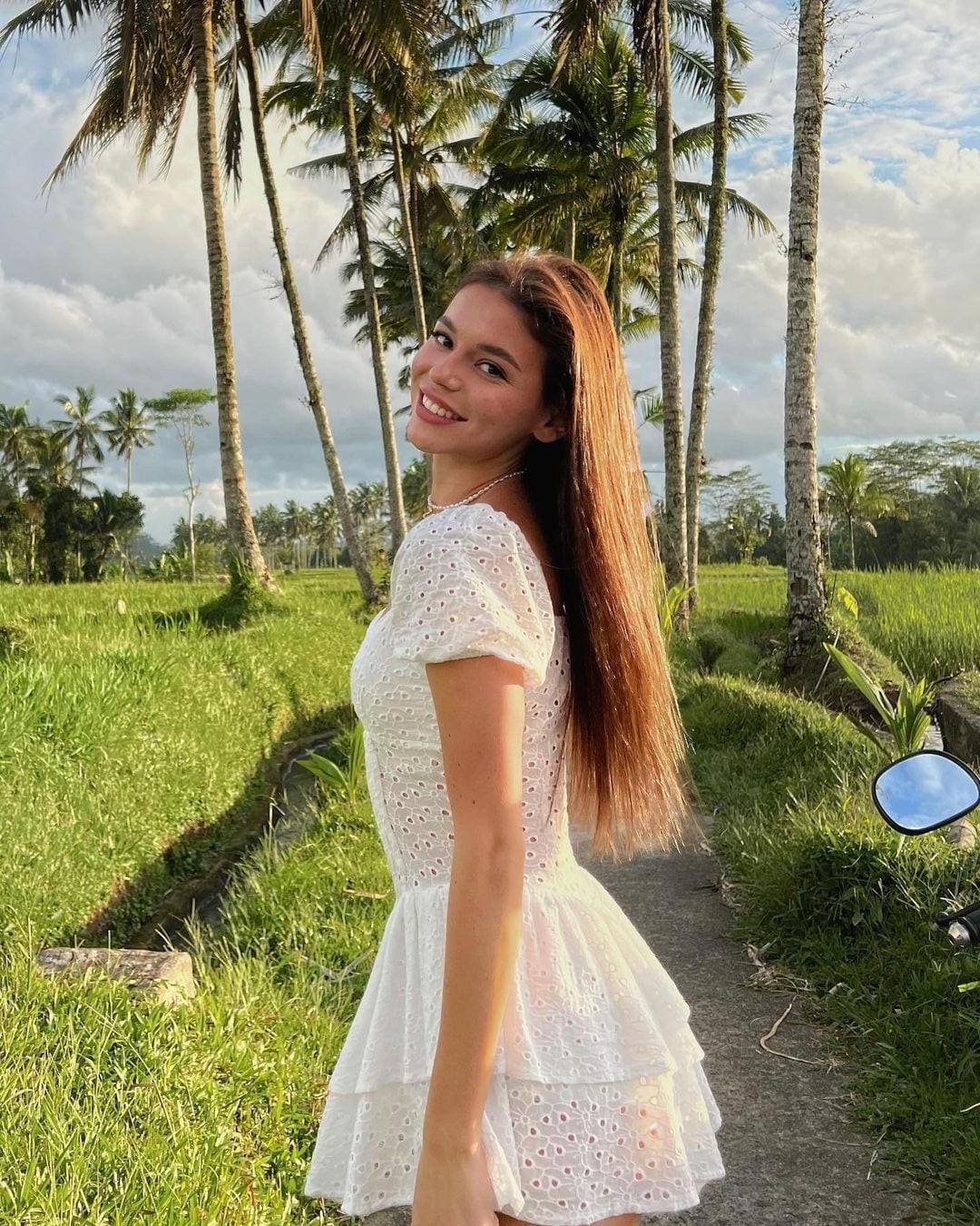 A woman in a short white eyelet dress stands on a path between rice paddies, with palm trees and clouds behind.
