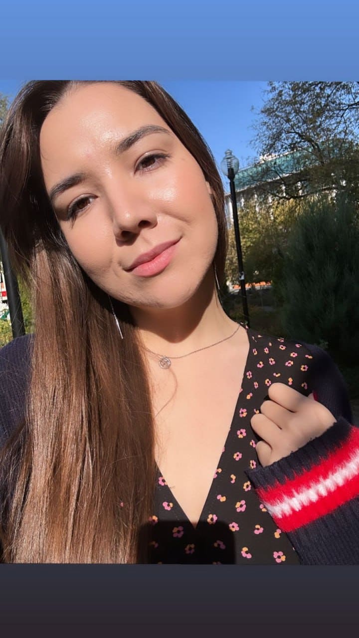 Moon takes an outdoor selfie against a blue sky, wearing a dark floral top with a striped sweater sleeve.