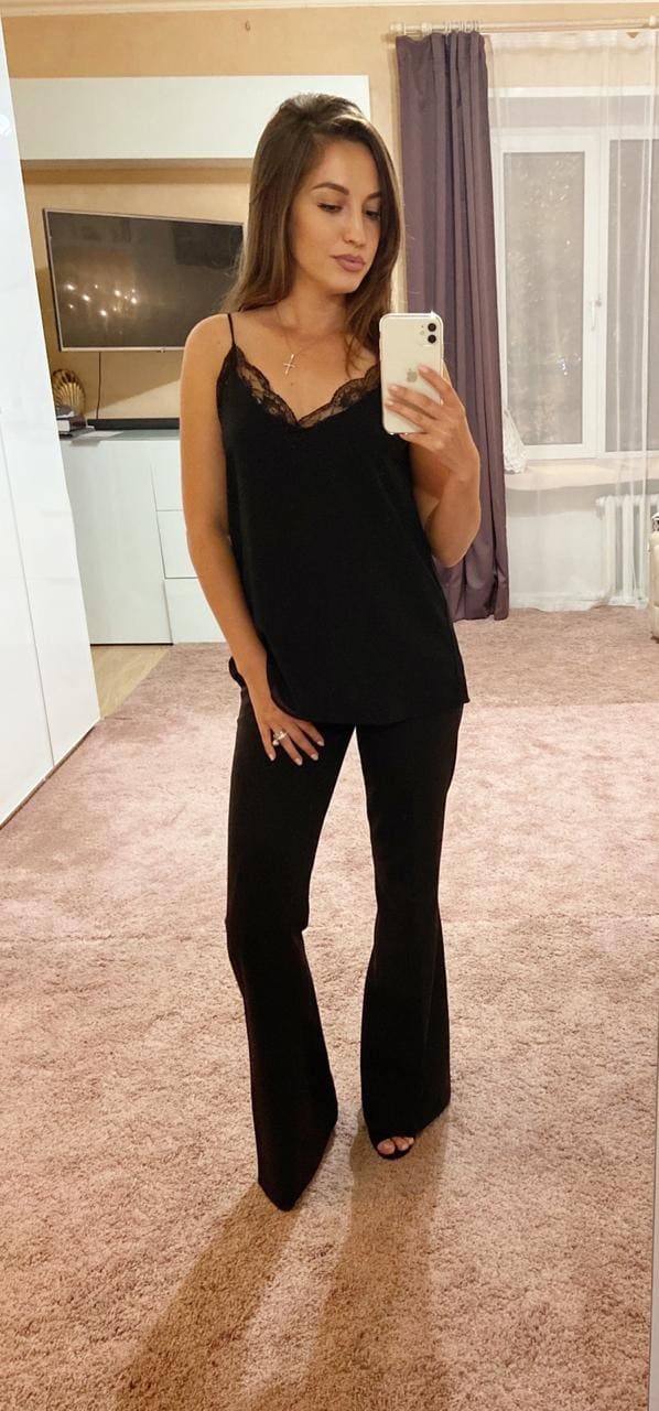 A mirror selfie shows a woman in a black lace camisole and flared trousers, holding a phone in a carpeted room with a TV.