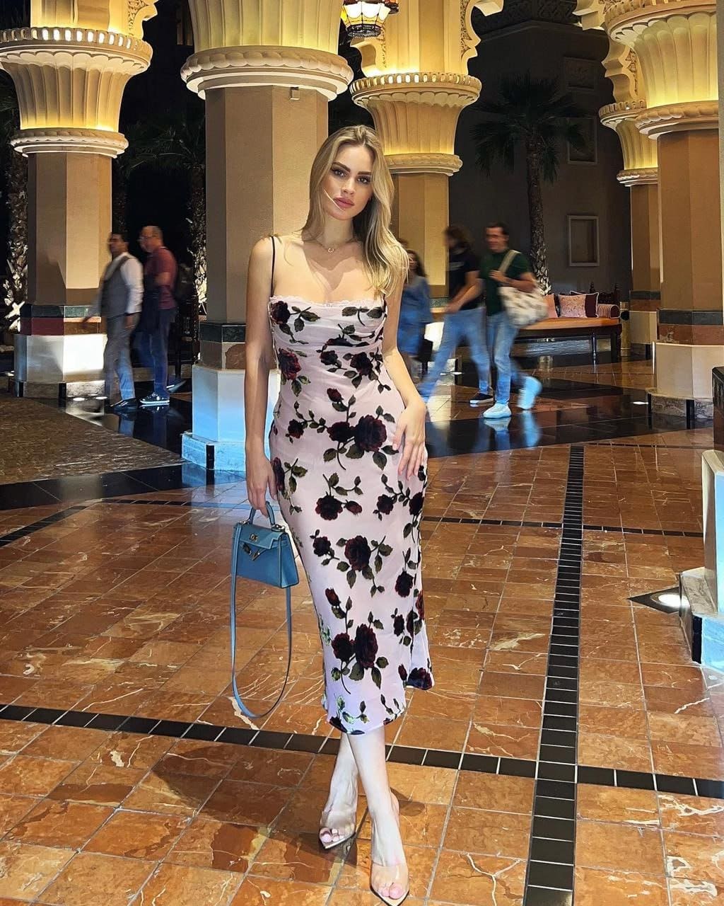 A blonde woman in a pink floral dress and clear heels stands in a tiled lobby with columns, holding a blue handbag.
