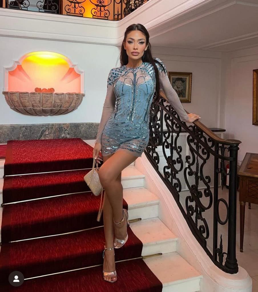 On a red staircase, she wears a sequined blue dress with sheer sleeves and heels.