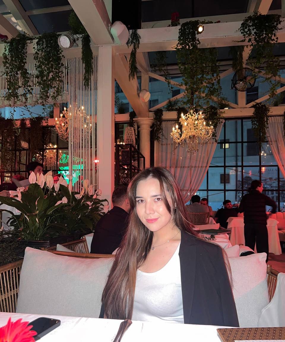 Woman in a white top and black blazer sits at a restaurant table beneath chandeliers and hanging plants.