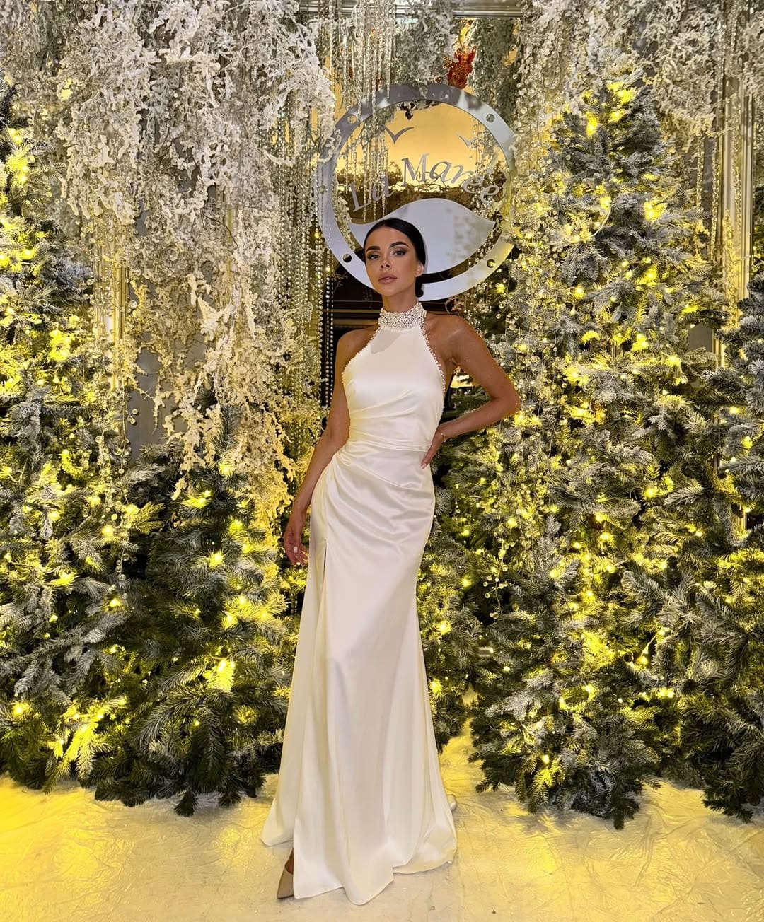 A woman in a floor-length white satin gown with a jeweled neckline stands with a hand on her hip before lit evergreen trees.