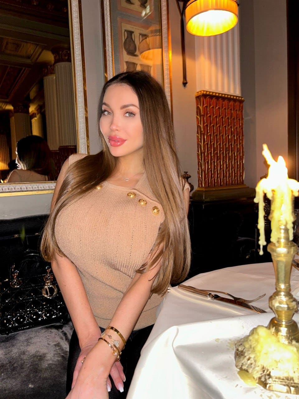Seated at a table in an ornate room, she wears a beige knit top with gold buttons and dark pants, beside a lit candle and black handbag.