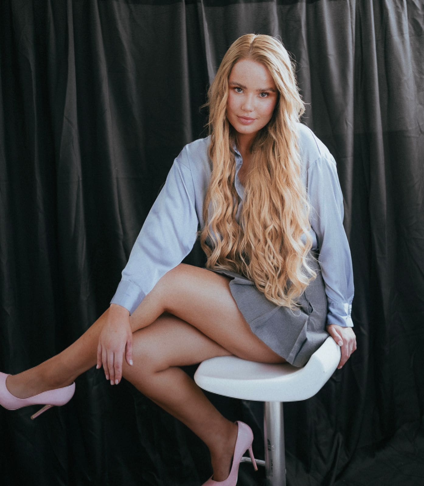 A woman sits on a white stool in an oversized light blue shirt, grey skirt, and pink heels, legs crossed before a black curtain.