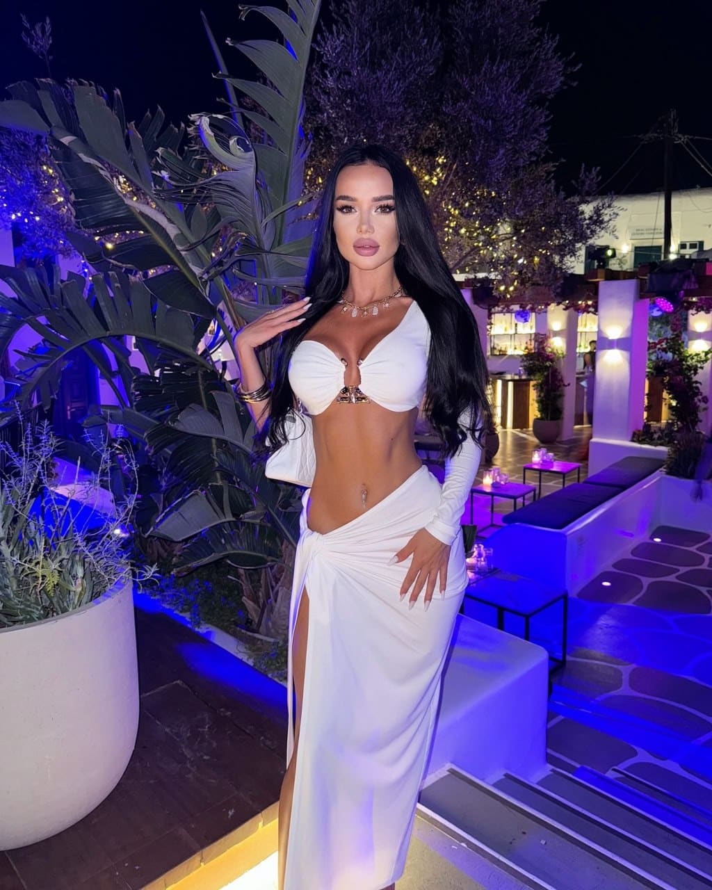 A woman stands on a night patio in a white one-shoulder crop top and high-slit wrap skirt, posing with one hand on her hip amidst purple lighting.