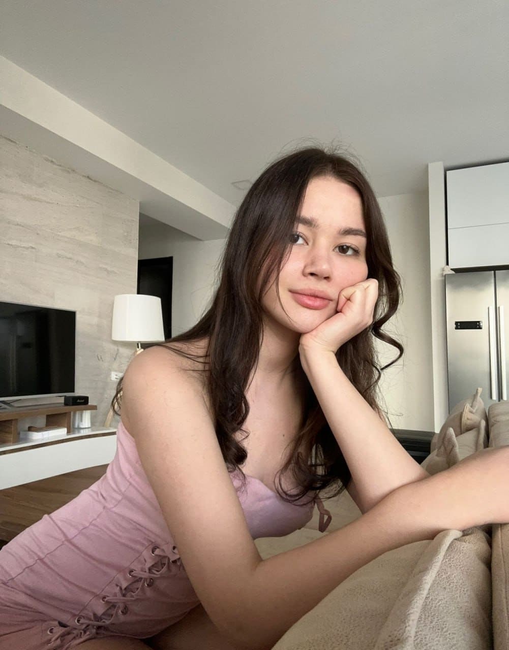 A woman sits on a beige couch in a modern living room, wearing a sleeveless pink lace-up dress and resting her chin on her hand.