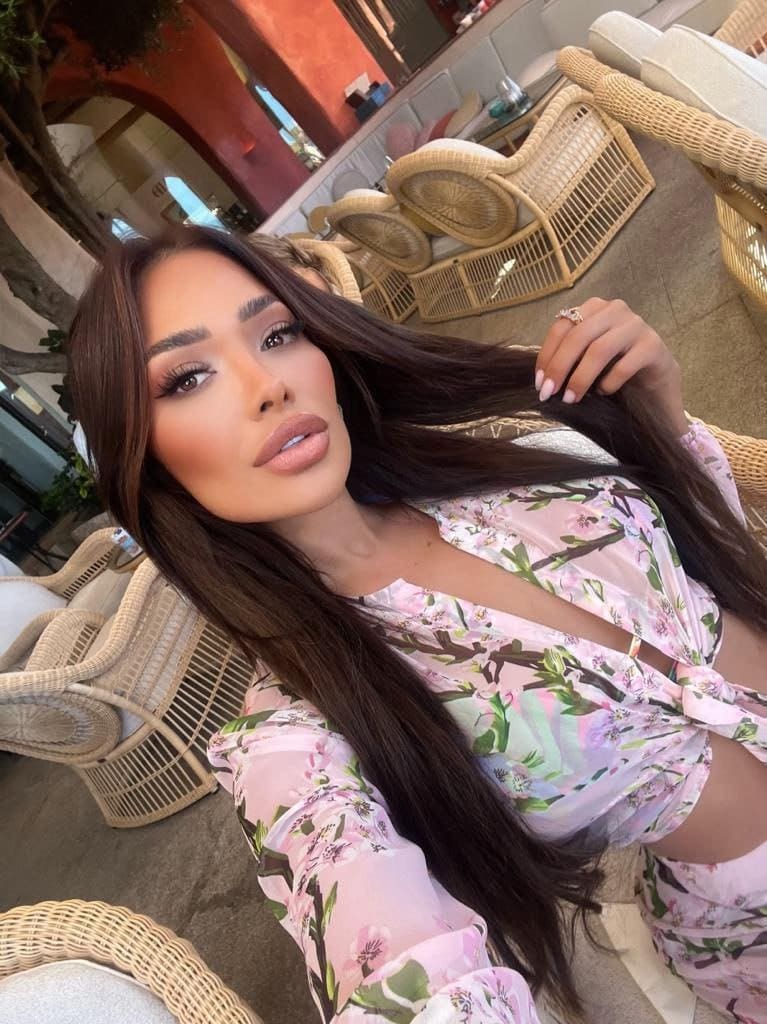 Seated in a wicker chair, she wears a pink floral two-piece outfit with cut-outs.