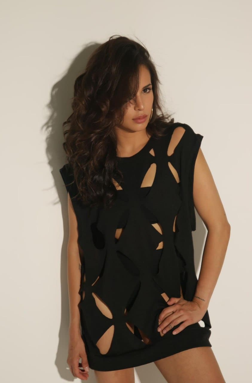 A woman stands against a white wall in a short black sleeveless dress with irregular cut-outs, one hand resting on her hip.