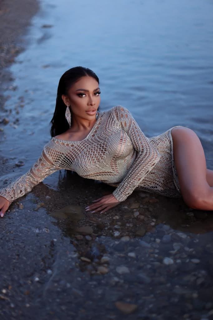 Lying on a wet shore, she wears an open-knit beige dress with a high slit.