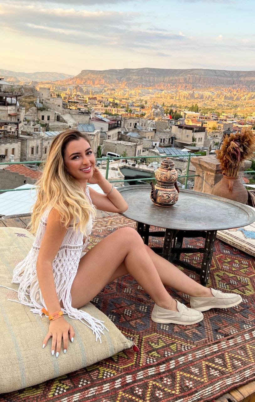 A blonde woman in a white fringed dress sits on a rooftop terrace with a canyon landscape behind her.