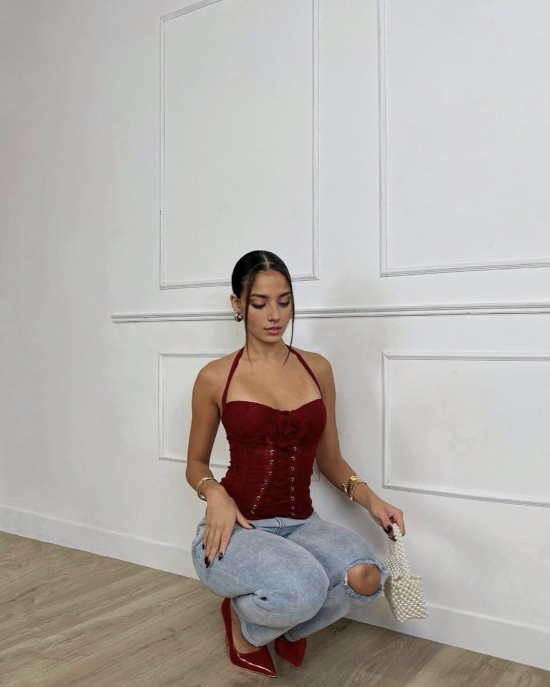 A woman in a red halter corset, ripped jeans, and red heels crouches on a wood floor, holding a small white beaded handbag.