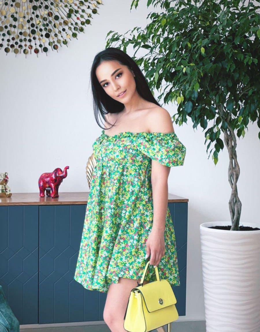 A woman with long dark hair wears a green floral off-the-shoulder dress and holds a yellow handbag in front of a blue cabinet.