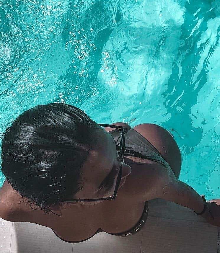 A person with wet hair and glasses sits at a turquoise pool edge in a dark bikini top, seen from above and behind.