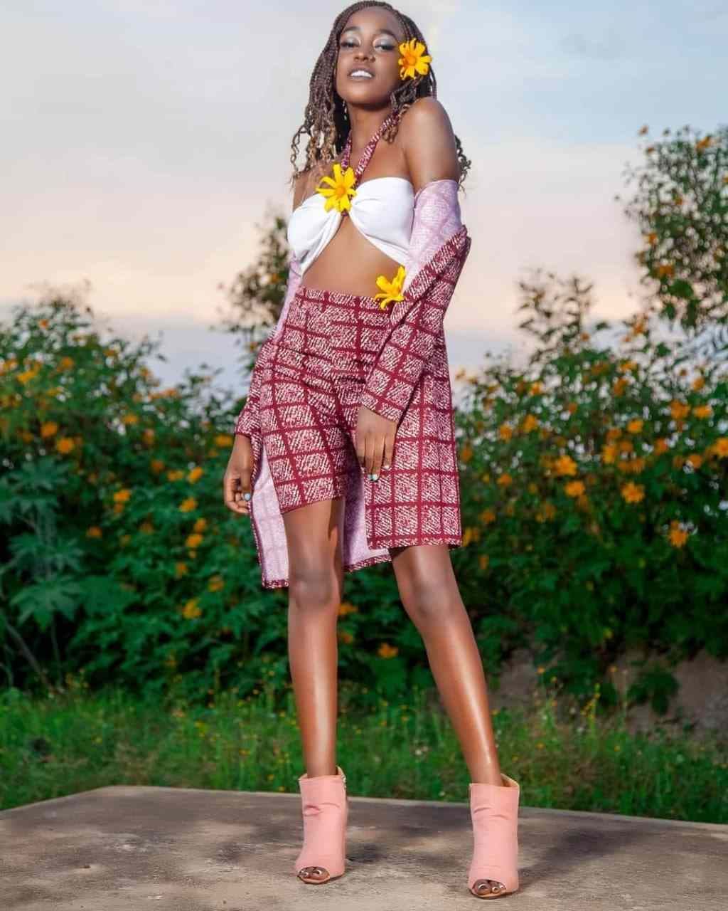 A woman with braided hair stands outdoors in a white bandeau, patterned wrap skirt, and pink boots against green foliage.