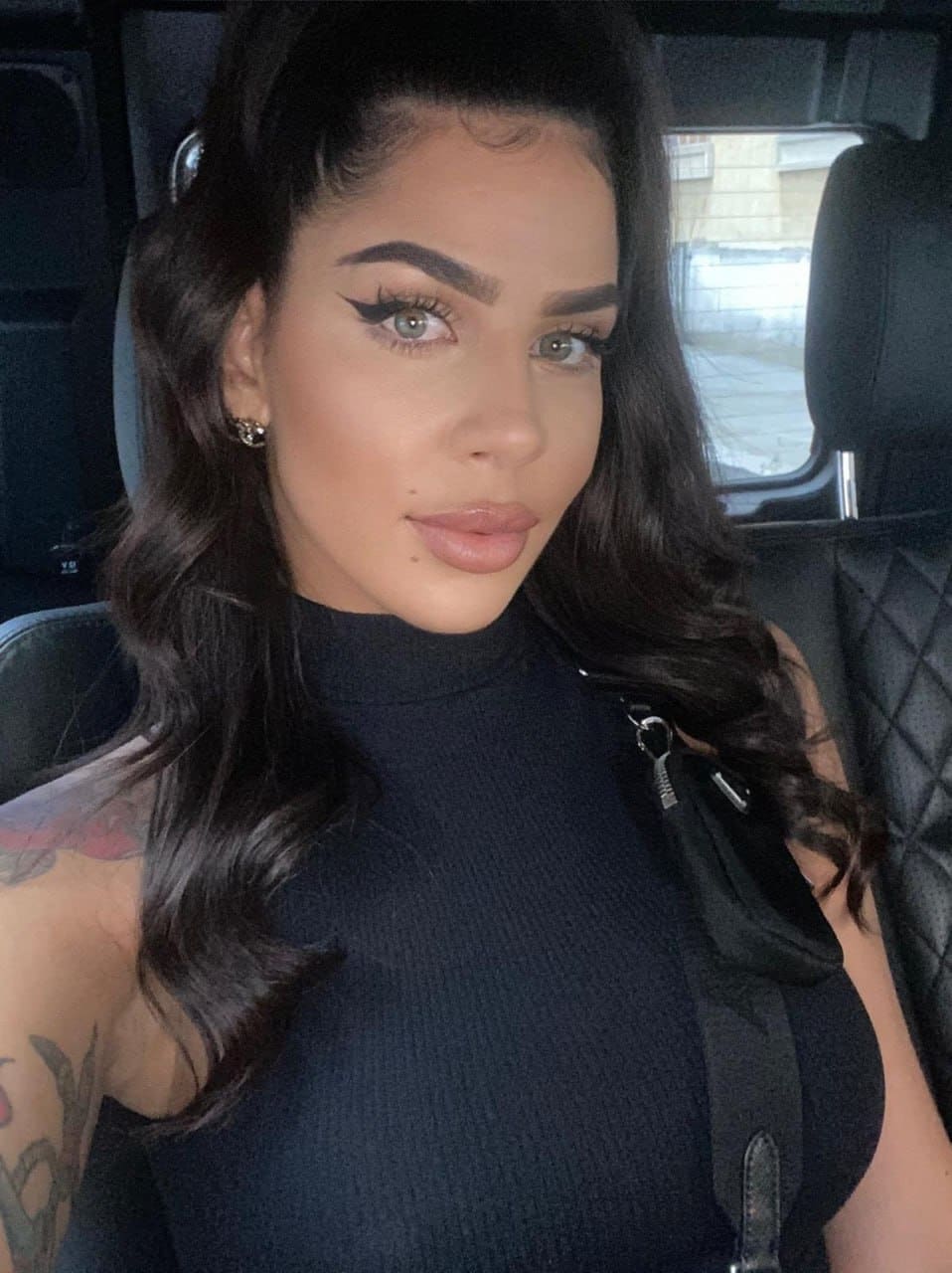 A woman with a shoulder tattoo sits in a car back seat wearing a black ribbed high-neck sleeveless top with a side zipper detail.