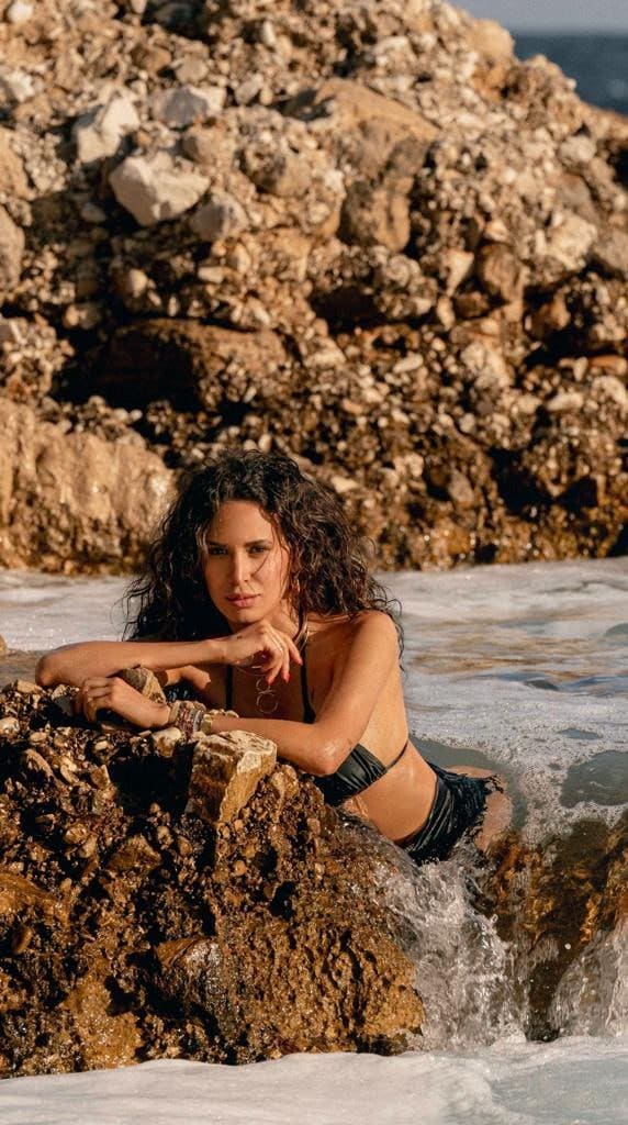 A woman reclines on a rocky shoreline in a black two-piece swimsuit, arms resting on rocks, with waves washing around her legs.