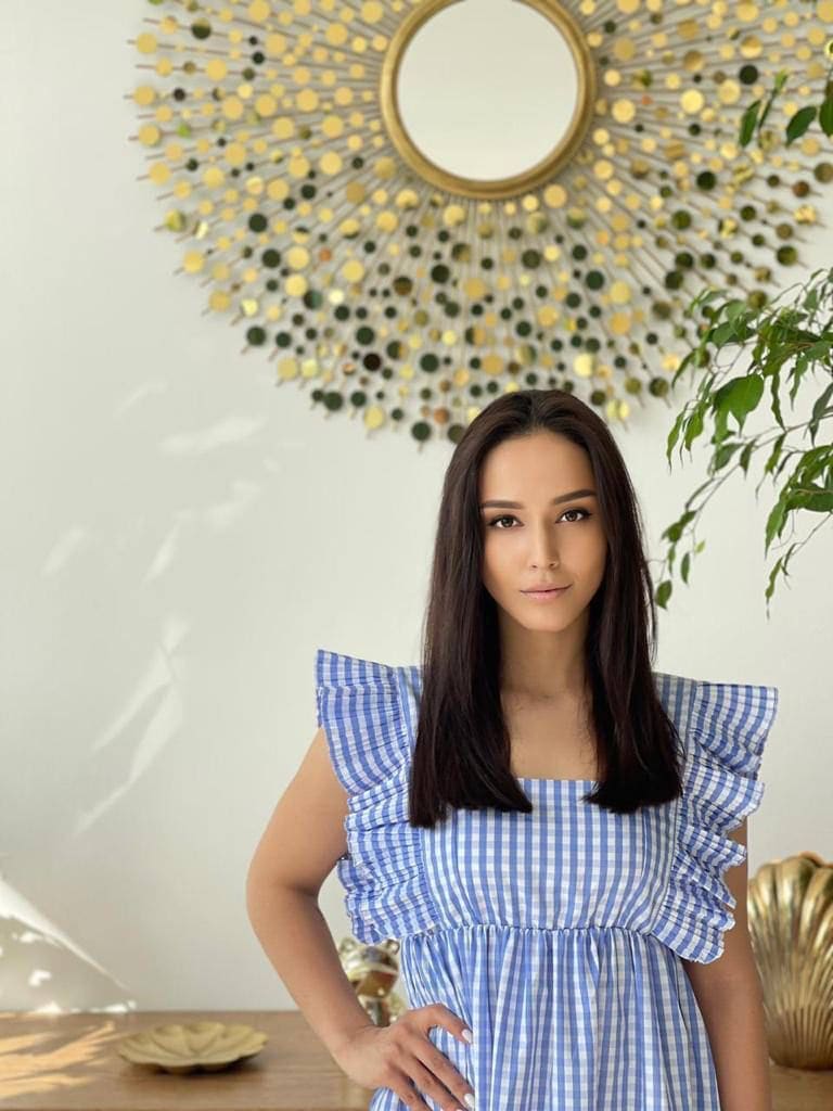 Lina stands with a hand on her hip in a blue and white gingham dress before a white wall with a gold sunburst mirror and a green plant.