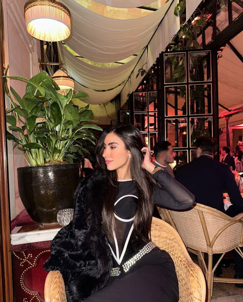 A woman sits in a wicker chair wearing a black top with white lines, a fur stole, and a beaded belt in a dimly lit restaurant.