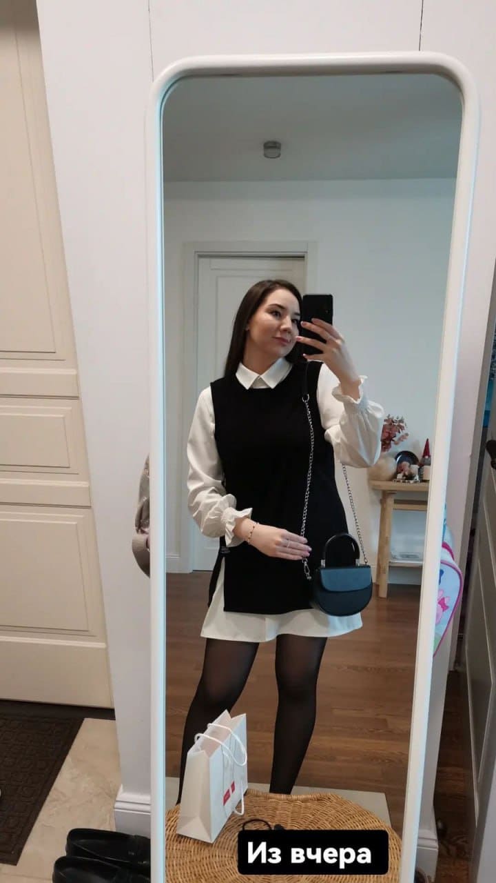Mirror selfie showing a woman in a white blouse, black sleeveless dress, tights, and chain-strap bag.