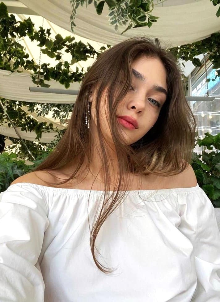 Woman in a white off-the-shoulder blouse tilts her head near hanging green plants and a white canopy.