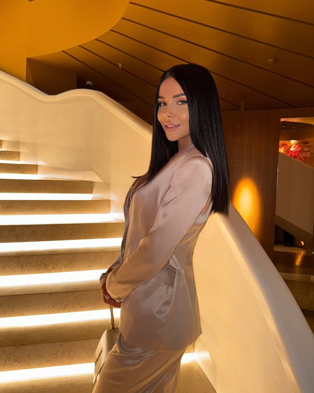 Margot stands in profile on a staircase with illuminated steps, wearing a beige satin blazer and skirt while holding a small handbag.