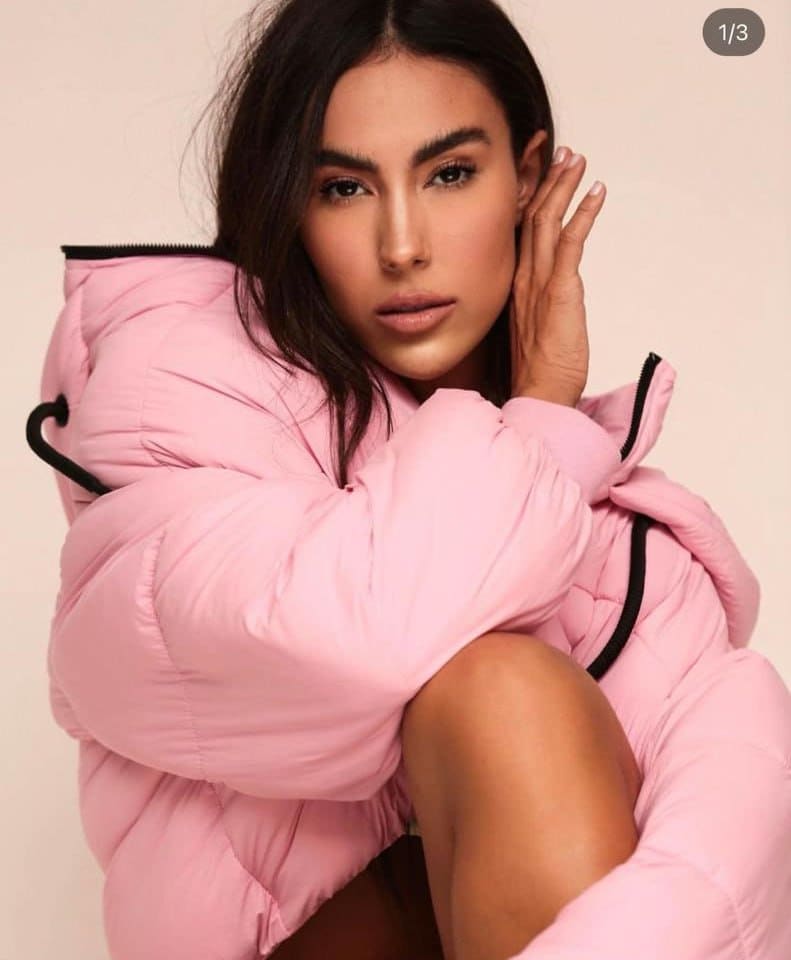 Ester sits with knees drawn up in an oversized pink puffer jacket, one hand raised to her head against a plain light background.
