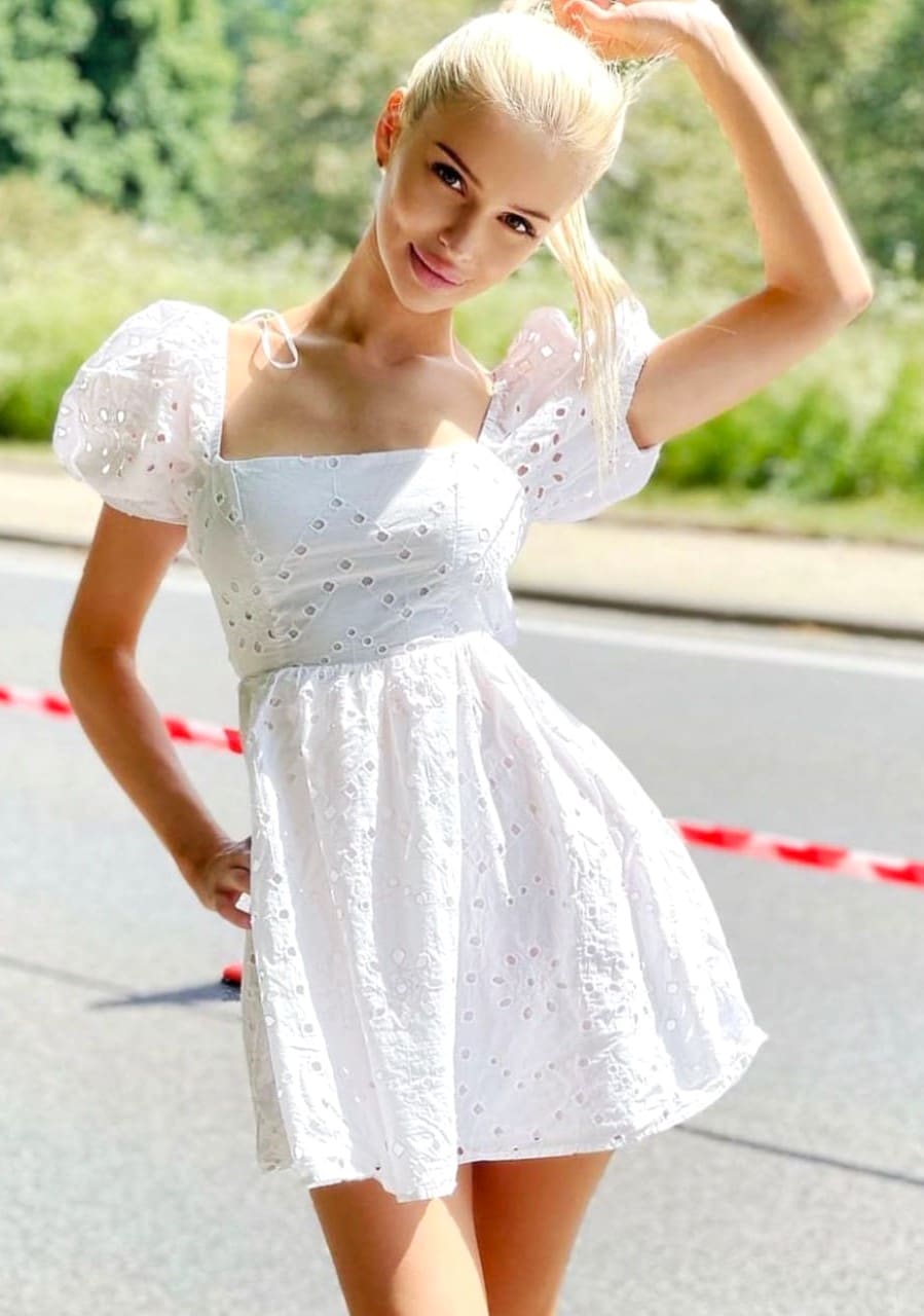 A woman in a short white eyelet dress stands on a paved road, one hand raised to her head, greenery behind.