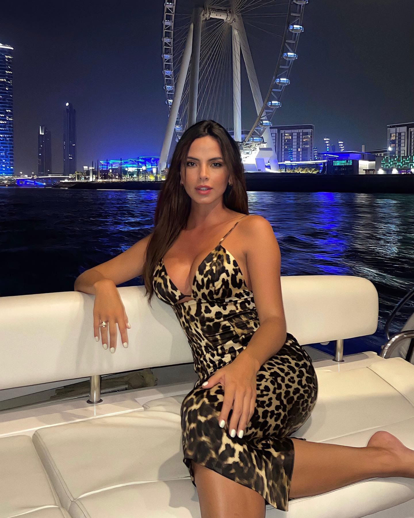 A woman in a leopard-print slip dress sits on white boat seating at night, with an illuminated Ferris wheel and city skyline behind.