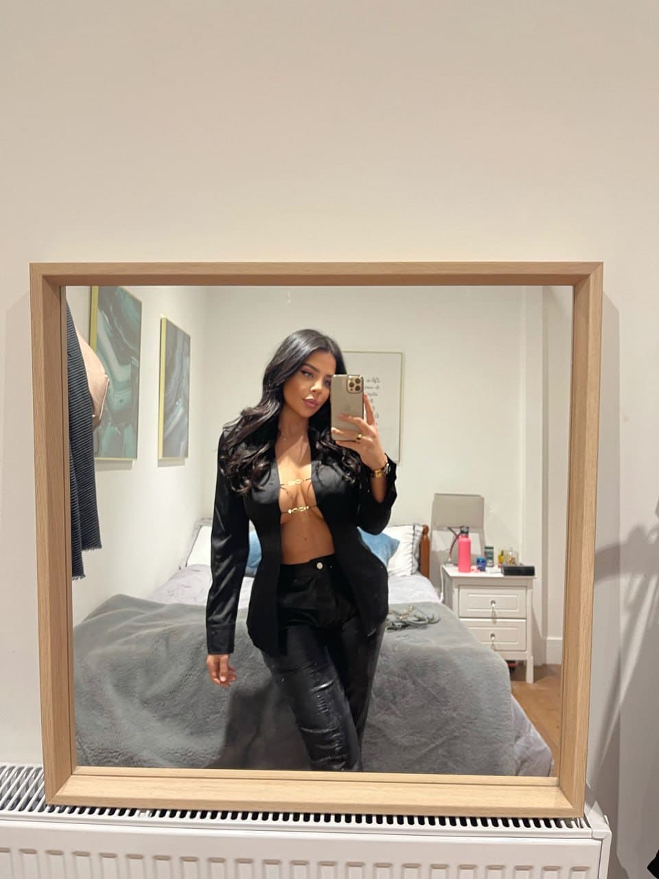A woman takes a mirror selfie in a bedroom wearing a black satin blazer over a gold bra and black leather pants, with a bed behind her.