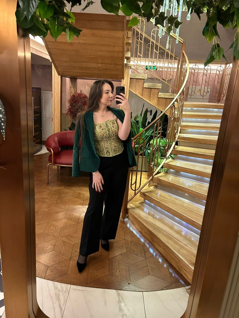 Woman in a green blazer and gold top takes a mirror selfie near a wooden spiral staircase and plants.