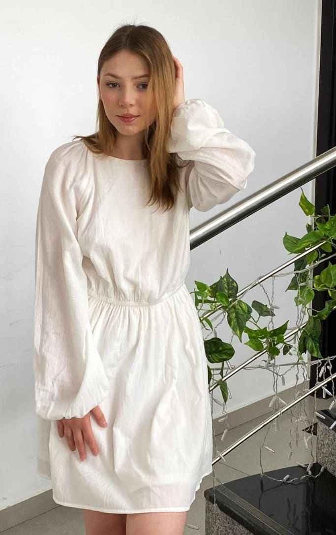 A woman in a short white dress with puffed sleeves stands indoors by a metal railing and plant, hand raised to head.